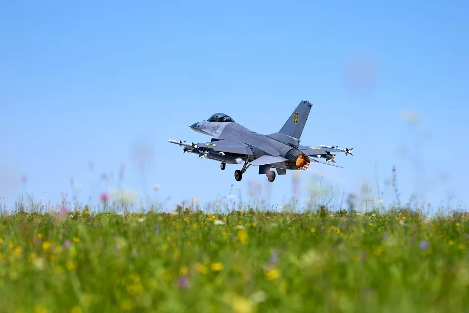 A view from ground level of a fighter jet taking off from a grassy field with flowers.