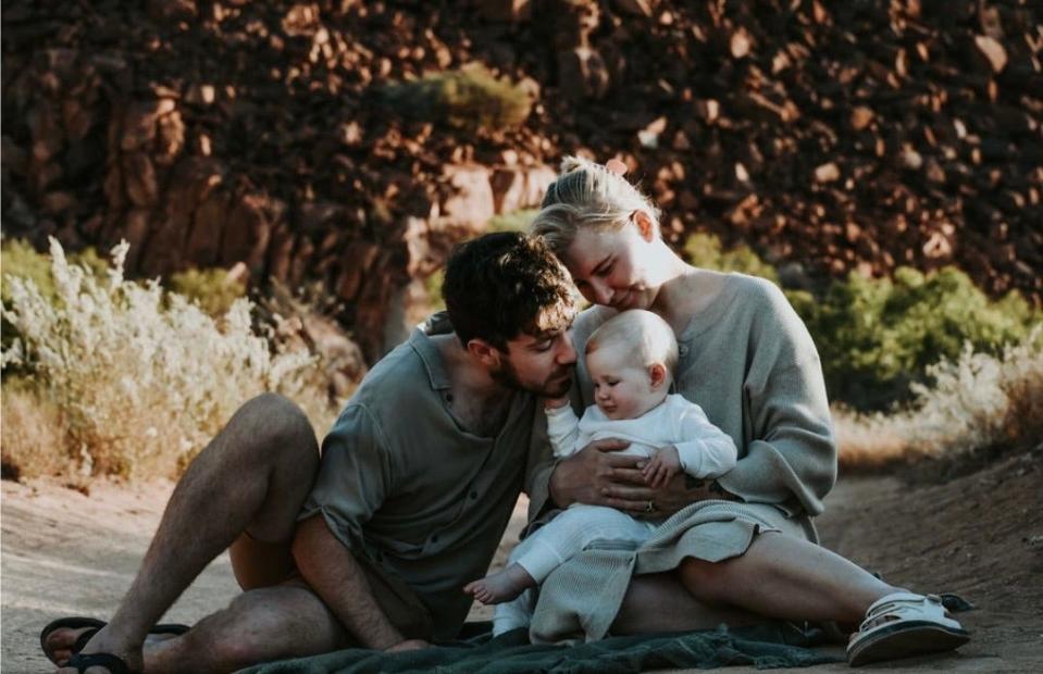 Megan Pagel with her husband and baby daughter sitting on a blanket on the ground outdoors.