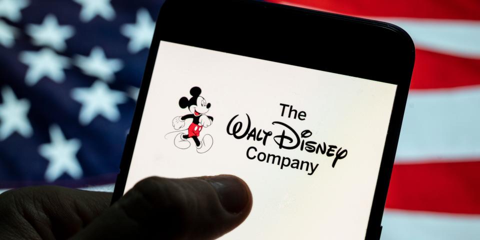 American flag in the background of someone holding a phone that says The Walt Disney Company on the screen.