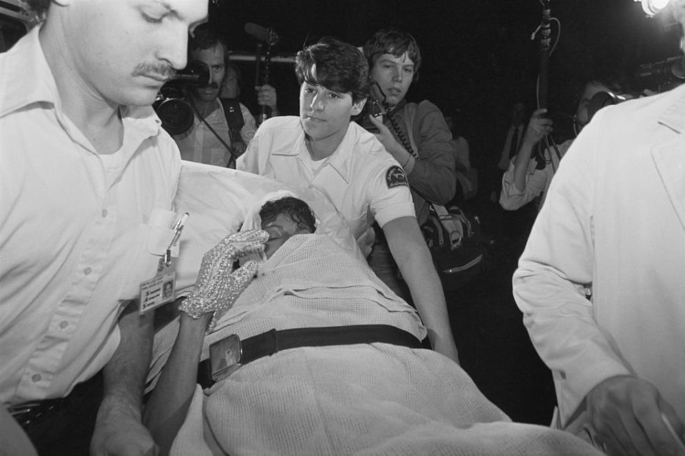 Michael Jackson being transported on a stretcher by emergency personnel, surrounded by press and photographers. Jackson's hand is adorned with a sparkling glove