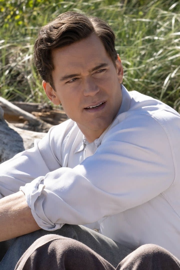 Matt Bomer and Jonathan Bailey sit on a sandy beach, both wearing white shirts and rolled-up pants. Bailey wears sunglasses and holds shoes in his hand