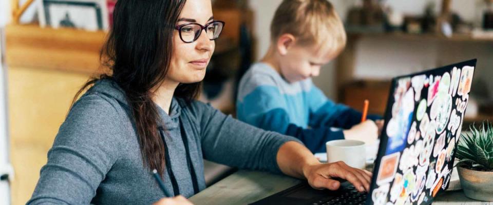 Concept of work from home and home family education. Mom and son are sitting at the desk. Business woman works on the Internet in a laptop, a child writes in a notebook.