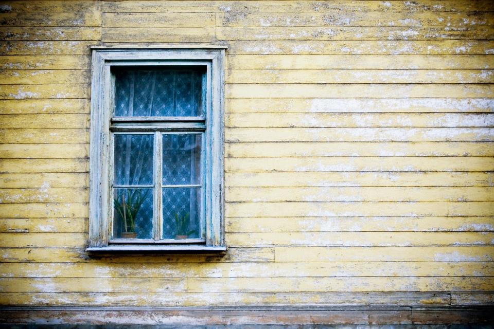 exterior wall of house with window with lace curtain and yellow siding that is faded and chipping