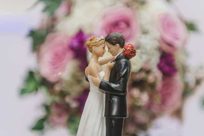 A bride and groom figurine stand in a romantic embrace in front of a blurred floral background. The bride wears a white dress and the groom a black suit