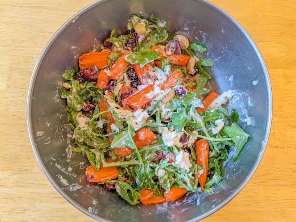 Bird's-eye-view of a metal bowl filled with arugula, carrot pieces, cranberries, nuts, and cheese. The ingredients are covered with a creamy-looking dressing.