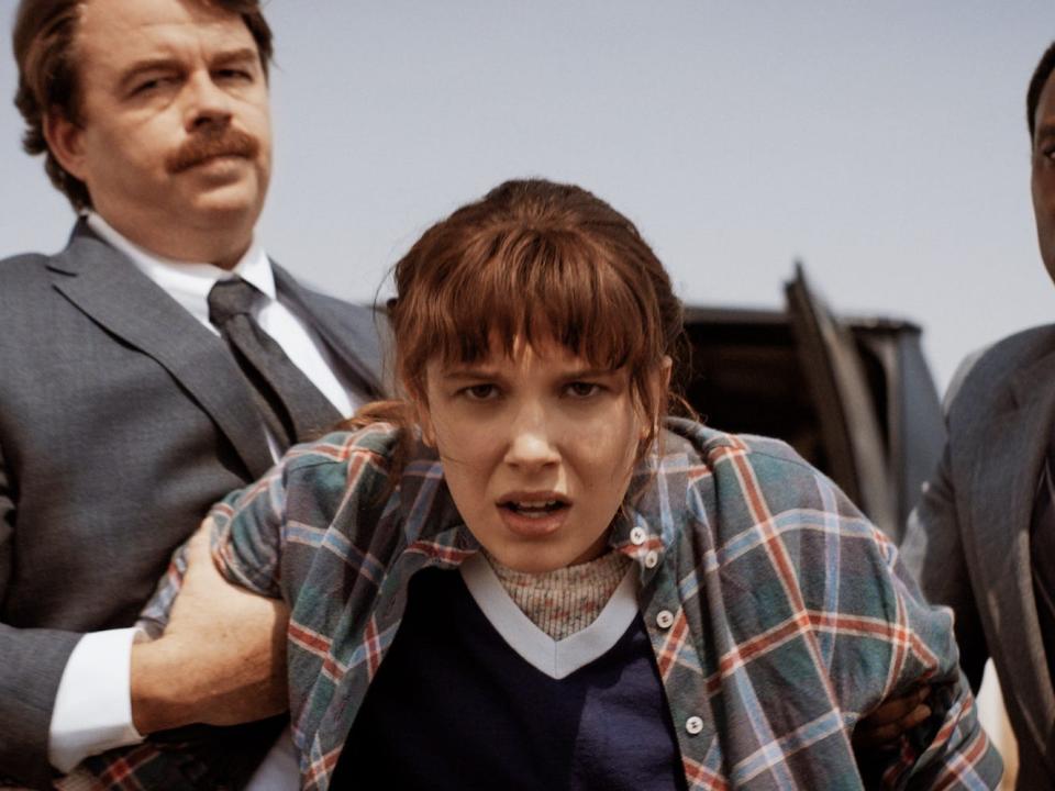 Two men in suits hold the arms of a teenage girl with brown hair, bangs, and wearing a plaid shirt (the character Eleven, played by Millie Bobby Brown).