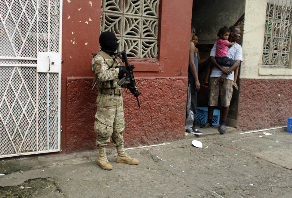 A masked police officer stands guard as residents look out from the doorway of their home in downtown Colon, Panama, Saturday, Oct. 20, 2012. Police began patrolling Colon after violent protests Friday over a new law allowing the sale of state-owned land to private companies already leasing land there to handle the import and export of goods in the duty-free zone next to the Panama Canal. Protesters want the government to instead raise the rent and invest the money in Colon, a poor and violent city. (AP Photo/Arnulfo Franco)