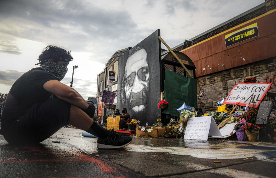 El manifestante Trevor Rodríguez observa sentado un monumento improvisado en el sitio donde George Floyd aparentemente murió después de ser sometido por la policía de Mineápolis y donde se pintó un mural y se han depositado flores y otros objetos en su memoria, el martes 2 de junio de 2020, en Mineápolis, Minnesota. (AP Foto/Bebeto Matthews)