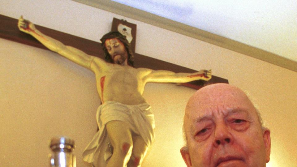 priest holding a vial of holy water in front of a crucifix statue