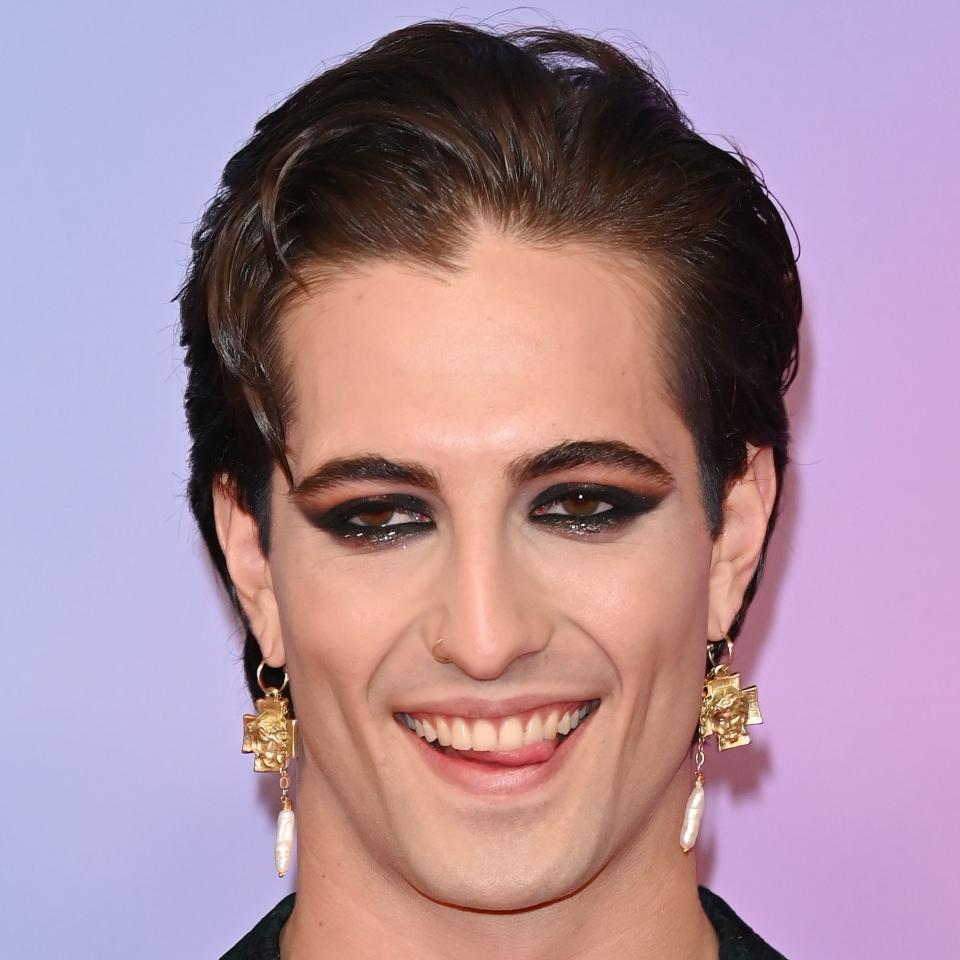 Damiano in a sheer top with bold makeup and earrings, smiling at an event