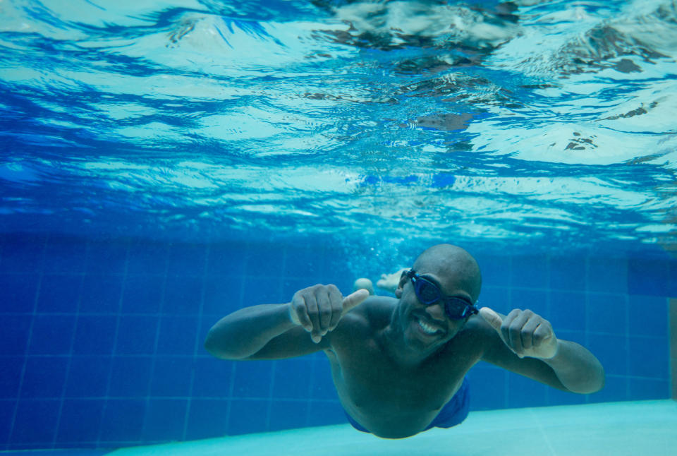 A person underwater in a swimming pool, wearing goggles and giving two thumbs up while smiling