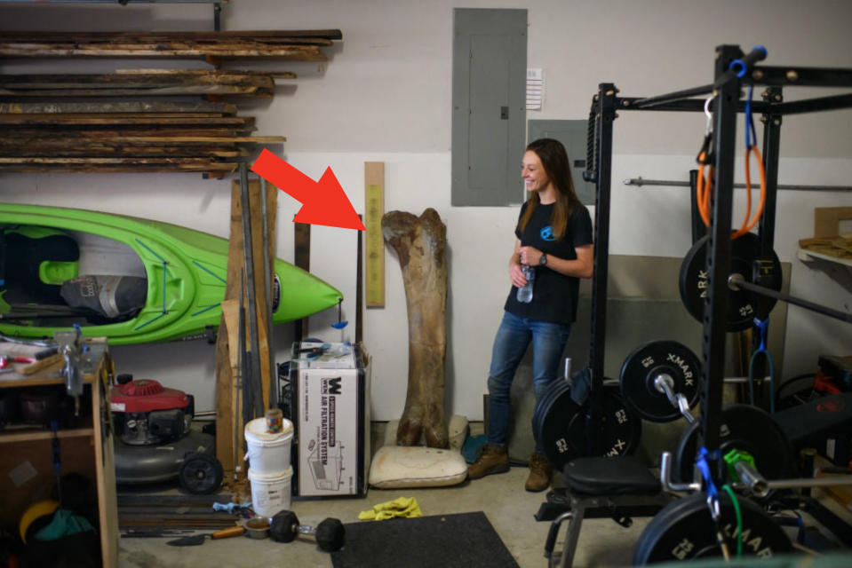 A woman stands in a garage filled with workout equipment, kayaks, and various tools. She is holding a water bottle and leaning against a wooden beam