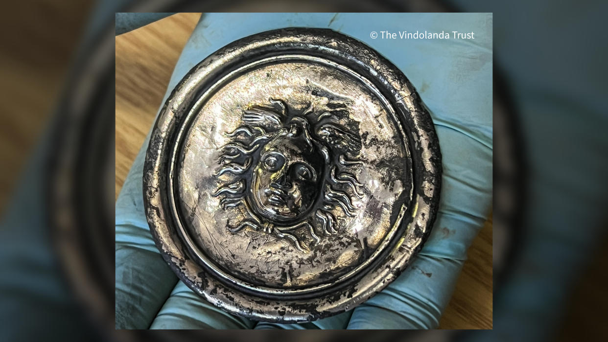  We see a circle-shaped silver disc with the face of medusa in the middle. A person wearing a blue glove is holding it. 