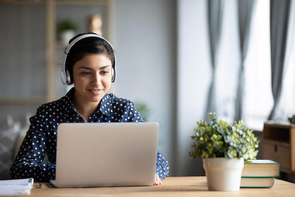 Millennial Indian girl in wireless headphones sit at desk at home working on modern laptop, young ethnic woman in earphones browsing Internet shopping online or studying on computer in living room