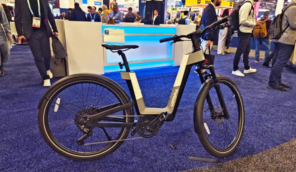یک دوچرخه برقی خاکستری، سفید و مشکی Urtopia Fusion بر روی پایه خود روی فرش آبی نمایشگاه CES 2024 قرار گرفته است.