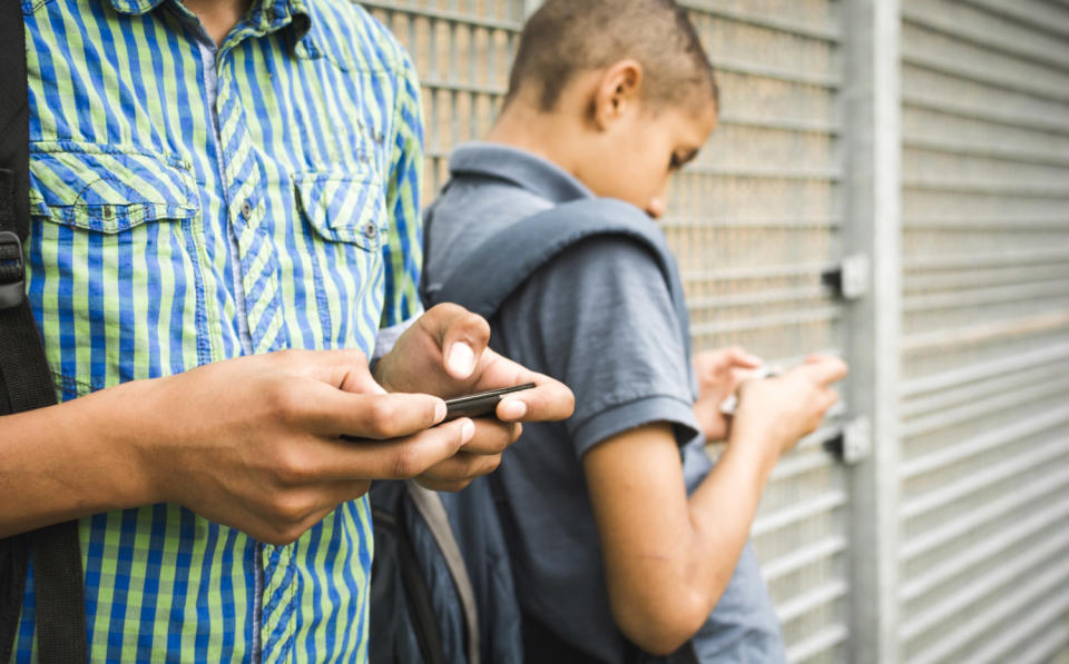 Two teenage boys leaning against a wall texting on smartphones.