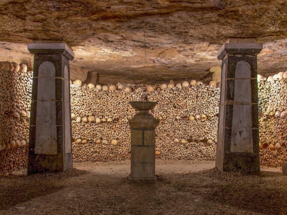 Catacombs with dim lighting and two pillars and a smaller pillar with a basin in the center of the room. The walls are lined with bones and skulls.