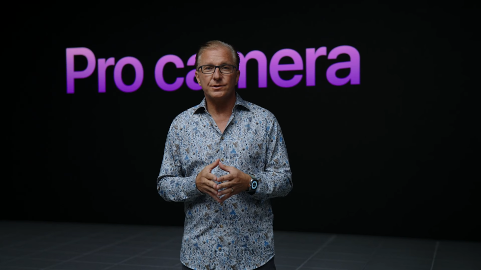 Still from the fall 2022 iPhone event. Apple VP Greg Joswiak stands in front of a dark background with purple lettering that reads, 