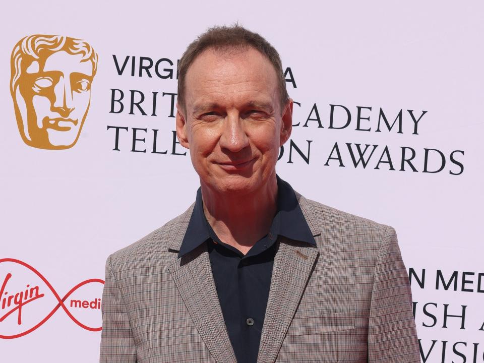 david thewlis wearing a grey suit with a black collared shirt, smiling on a red carpet and squinting his eyes slightly into the sun