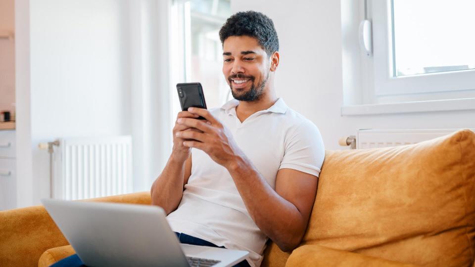 Smiling casual mixed-race freelancer using smart phone and laptop while sitting on the sofa.