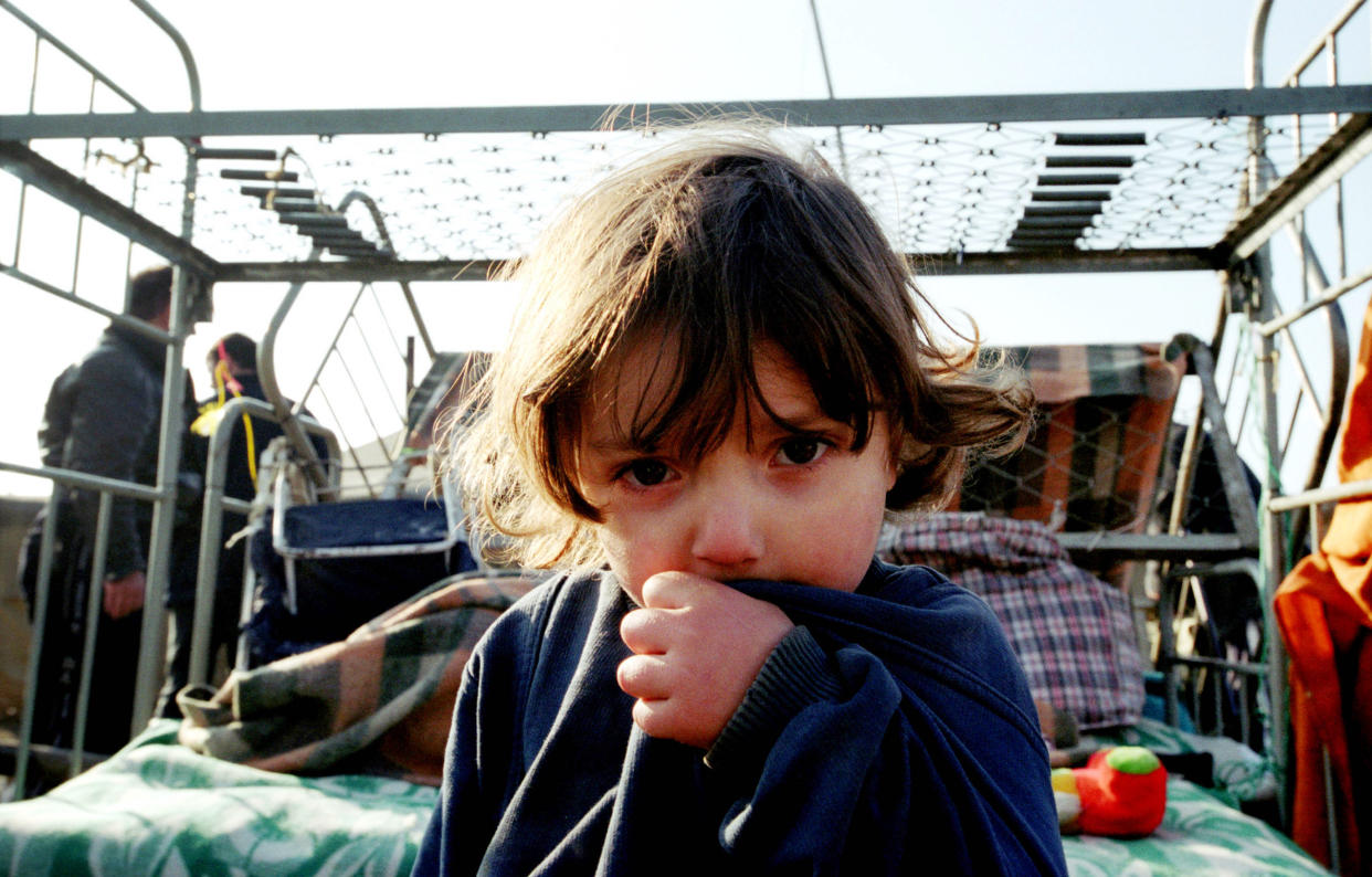 A young child sits on a bed, facing the camera, with a backdrop of refugees.