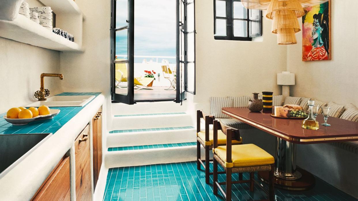 a kitchen with turquoise tiled floor and a wood table with yellow chairs and a banquette with striped fabric, rattan pendant, counter with sink and shelves above and wood cabinets below, steps go to a doorway out to terrace