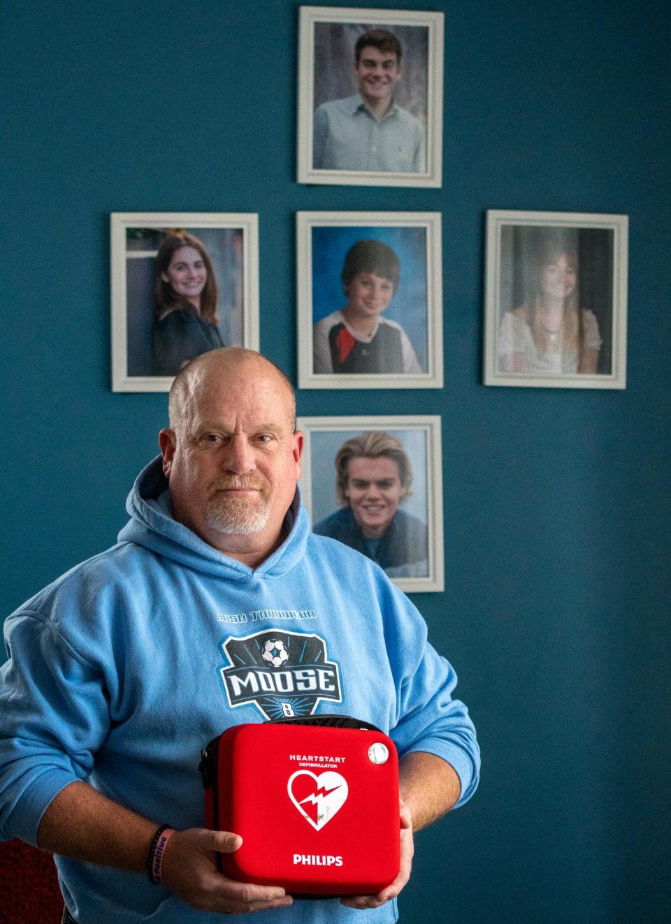Ralph Thibodeau holds a defibrillator by photos of his children, including Josh in the middle, Tuesday at his Holden home. Josh was 12 years old when he died unexpectedly at a soccer camp in 2011.