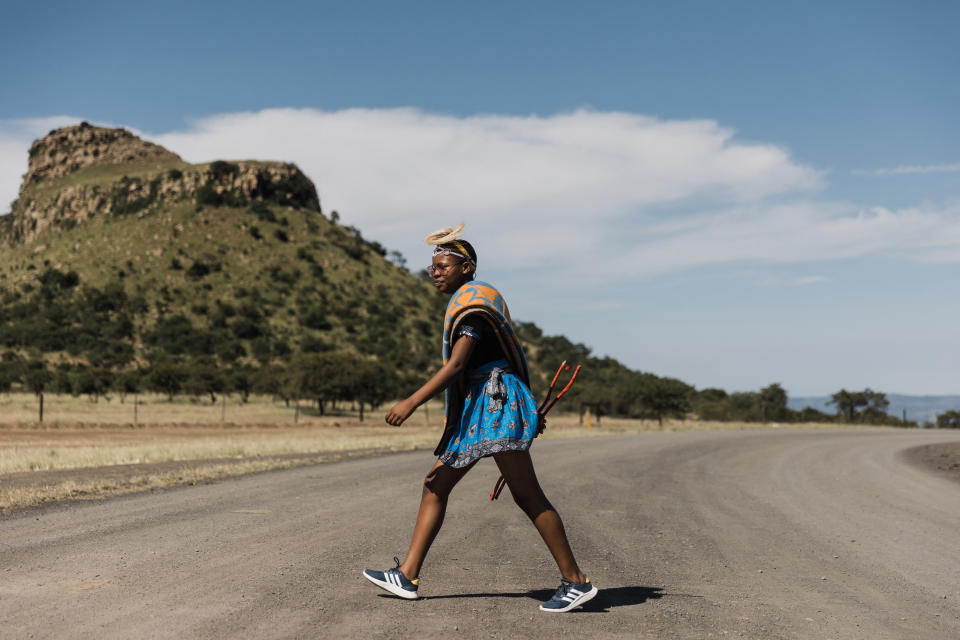Someone wearing traditional Basotho attire and Adidas tennis shoes walks across a road with a mountain in the background.