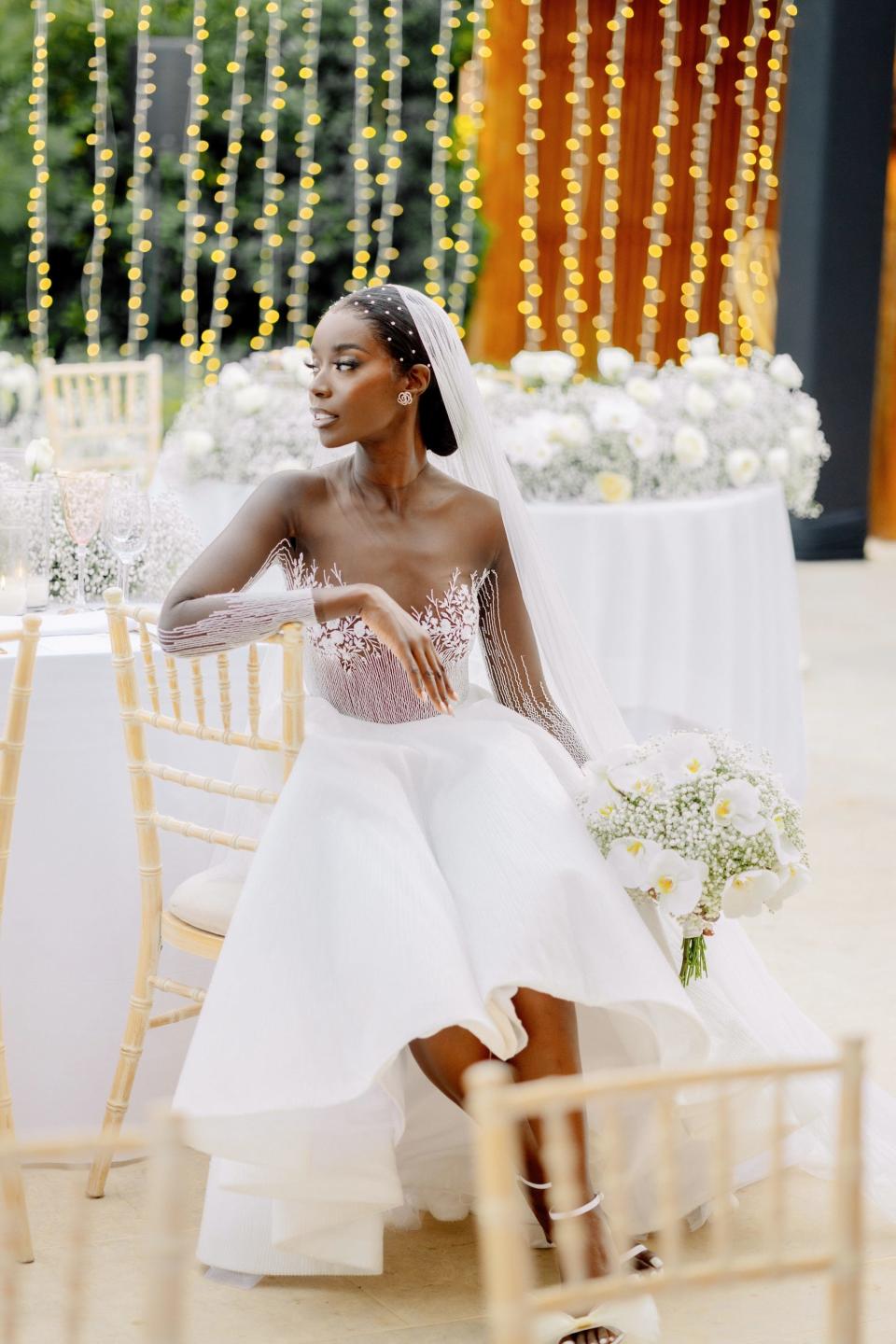 A bride sits in a chair in her wedding dress in front of a wall of twinkly lights.