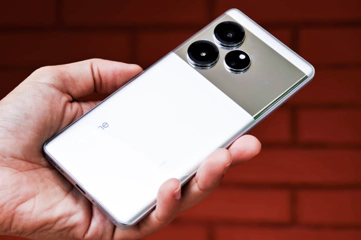 Realme GT 6 Android phone in silver color held in hand in front of a red brick wall.