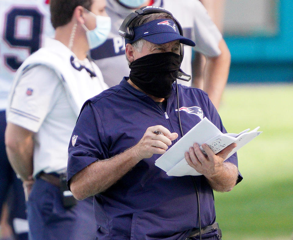 Bill Belichick wears a mask, visor and headset while he holds a stack of papers and looks on during the game against the Dolphins.