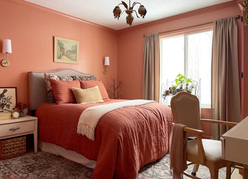 Neatly made bed with rust colored bedding in monochromatic bedroom. Arm chair tucked under writing desk