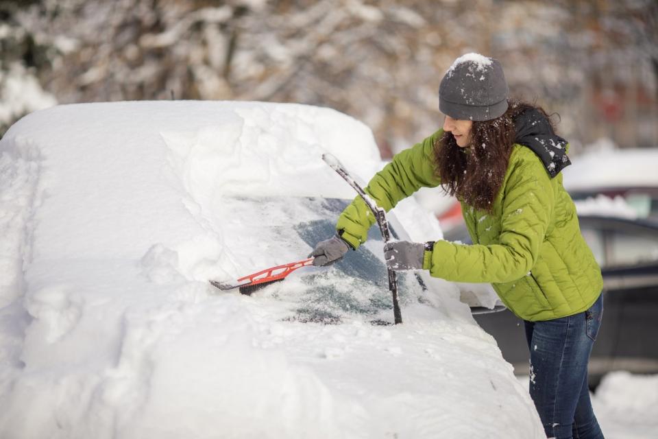 Woman cleaning her car from snow on parking. She wears warm clothes and uses a brush for snow.