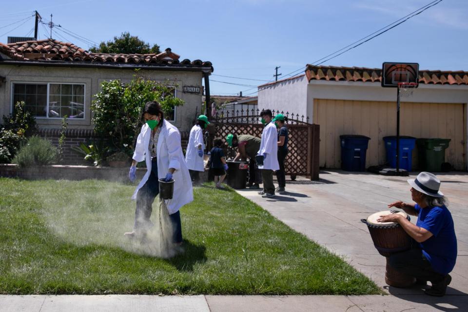 A woman in a mask and lab coat uses a small bucket to dust a home's frontyard with a powdered mineral.