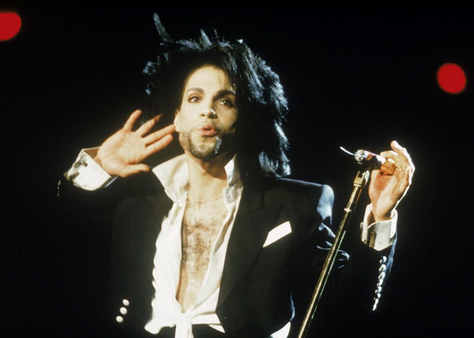 Prince stands on stage with a microphone, one hand by his ear to hear the crowd response, during a concert, February 1991. 