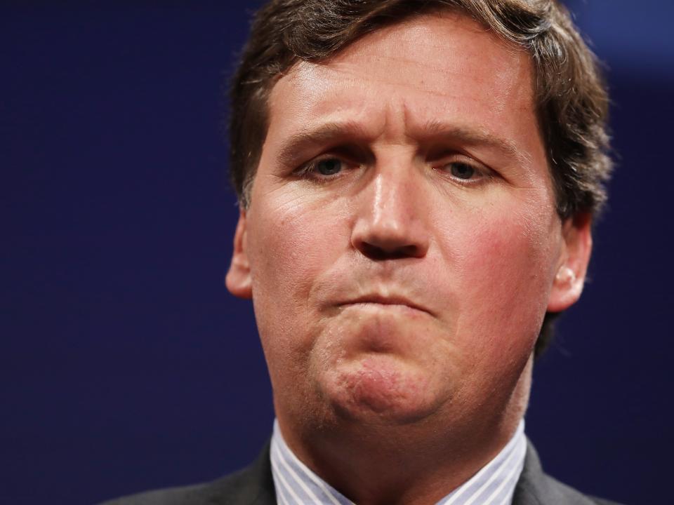 A headshot of Tucker Carlson looking concerned while standing in front of a blue background and wearing a suit and tie.