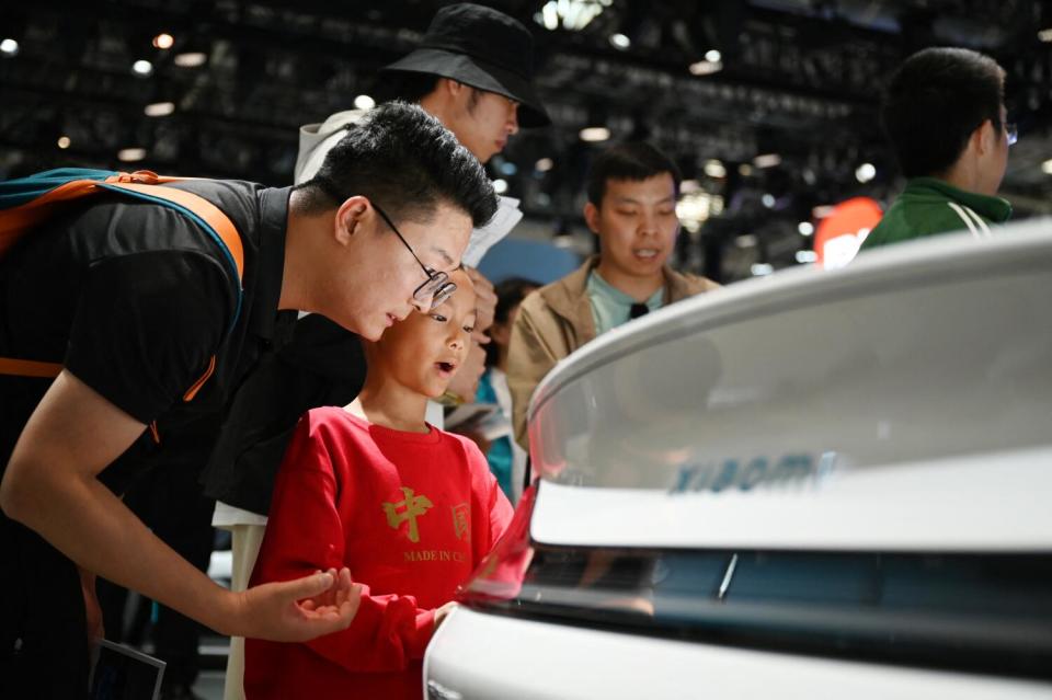 A person in a dark shirt and glasses leans down next to a child in a red top looking at a vehicle