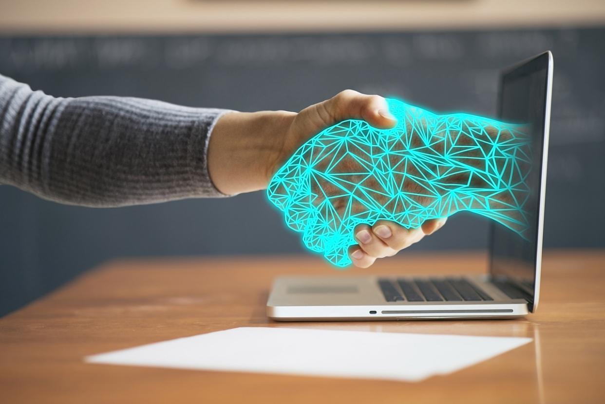  A human hand shakes a digital hand coming out of the screen of a laptop in this illustration of AI assisting a human. 