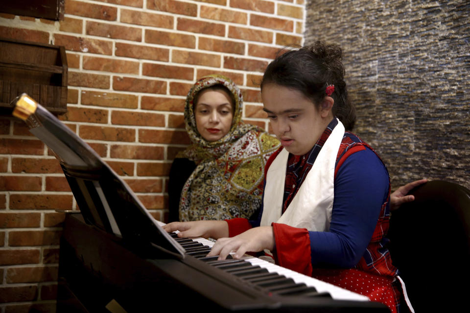 In this Monday, Aug. 6, 2018, photo, cafe staffer Melika Aghaei, 14, with down syndrome plays piano, while Aylin Agahi founder of Downtism Cafe and a musician listens, in Tehran, Iran. The popular cafe in Tehran’s bustling Vanak Square, whose name combines “Down” with “autism,” is entirely run by people with Down syndrome or autism. More than just providing meaningful work, the cafe is helping break down barriers by highlighting how capable people with disabilities are. (AP Photo/Ebrahim Noroozi)