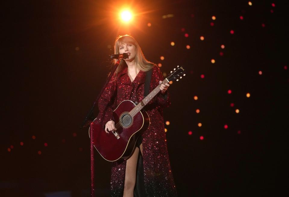 Taylor Swift singing into a microphone on stage while playing a red acoustic guitar