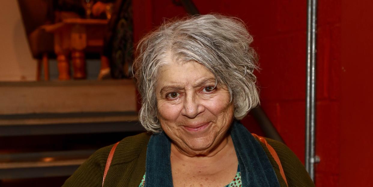 miriam margolyes in 2019, posing in front of a red wall, she is wearing a green cardigan and scarf and green and red dress and has short grey hair