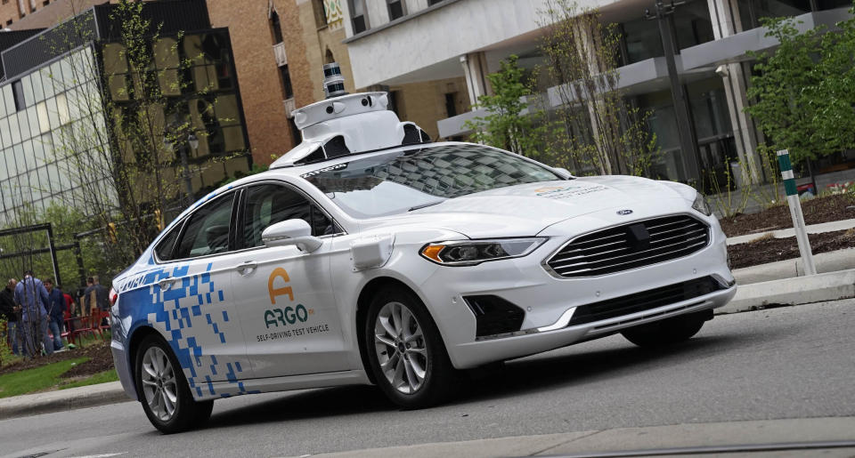 A white Ford Fusion sedan with Argo AI logos and visible self-driving sensor hardware is shown driving on a Detroit city street.