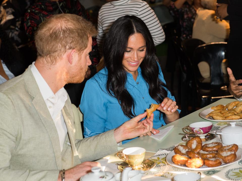 Harry and Meghan sitting at a table with food in front of them and splitting and pulling apart a samosa pastry to share with their hand.s