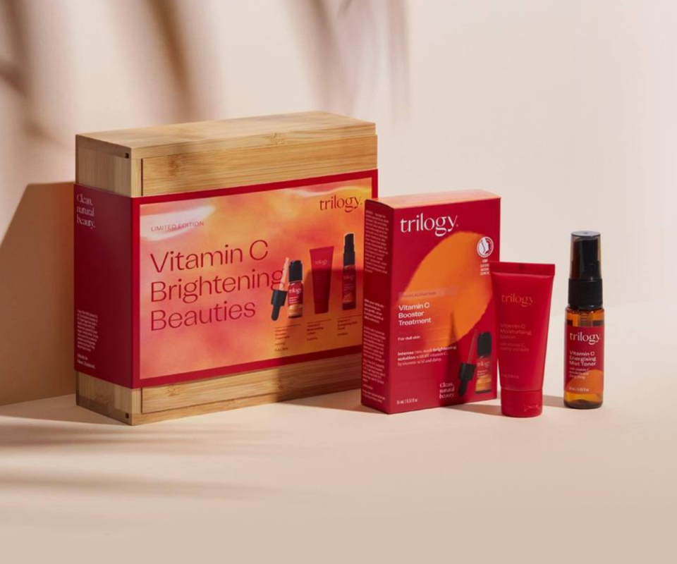 Trilogy's Vitamin-C skincare set in a wooden box sits upright infront of a pink wall, with the products in red packaging infront of it along with the brown oil bottle and red moisturiser infront of it. 