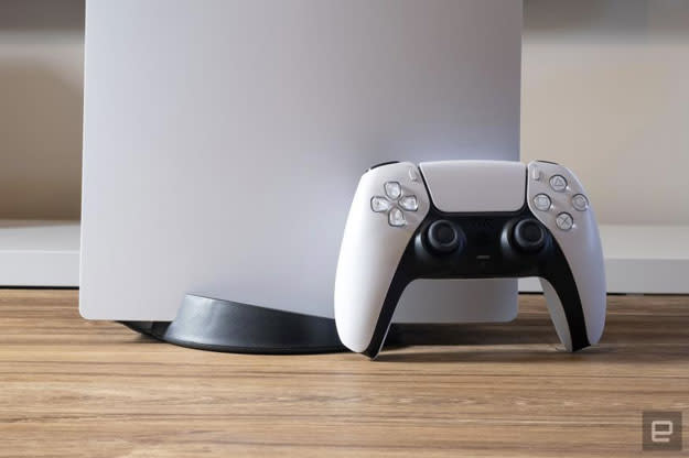 Image of the lower half of a PlayStation 5 and a single controller propped up against it on a wooden table. 