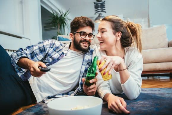 Couple drinking beer while lounging on the floor with a bowl of popcorn between them.