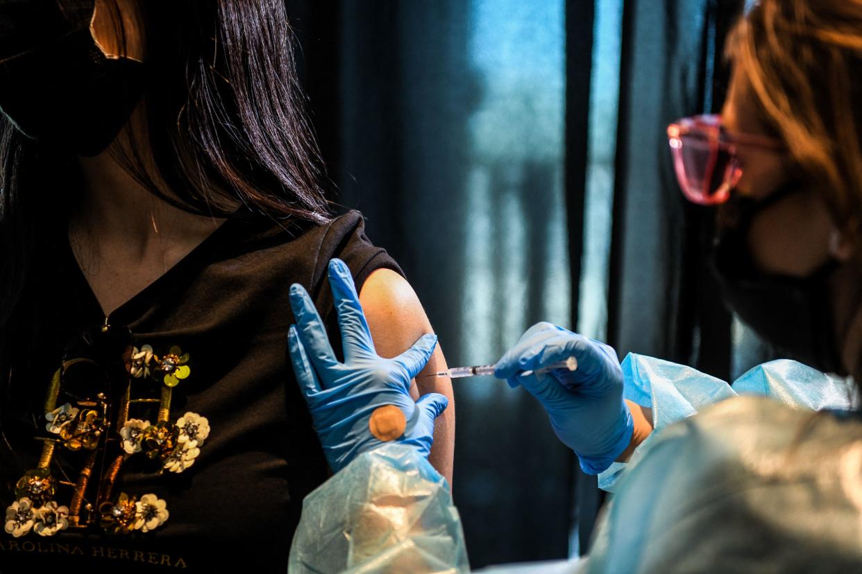 A health care worker wearing blue rubber gloves, a face mask and protective clothing administers a shot to the upper arm of a young woman.