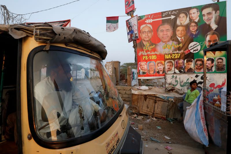 A rickshaw (tuk tuk) driver waits for a customer, as his vehicle is parked beside a campaign poster of a political party, ahead of general elections, in Karachi