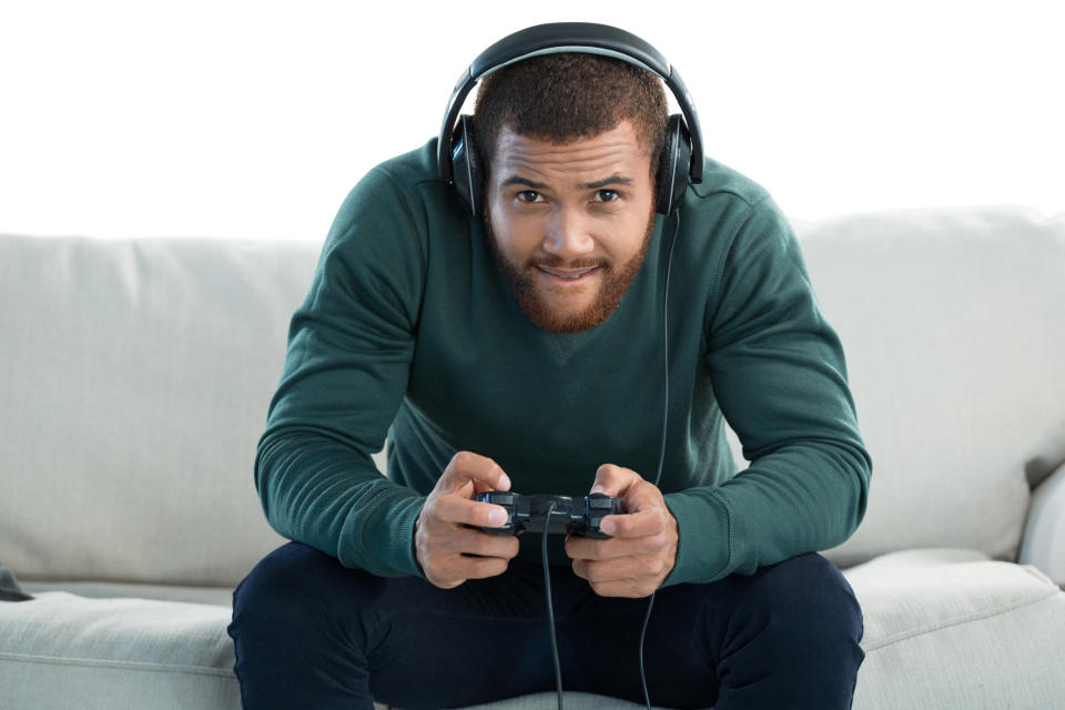 Man in green sweater and black pants sitting on a white couch. He's leaning into the TV screen, wearing headphones and wielding a video game controller.