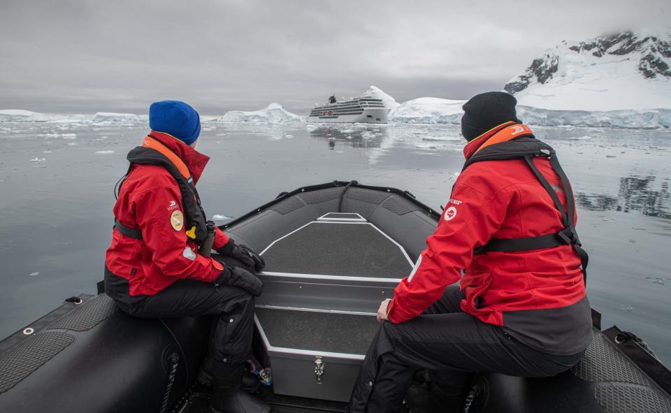 Two people in red jackets and hats sit in a zodiac boat looking at a cruise ship in Antarctica